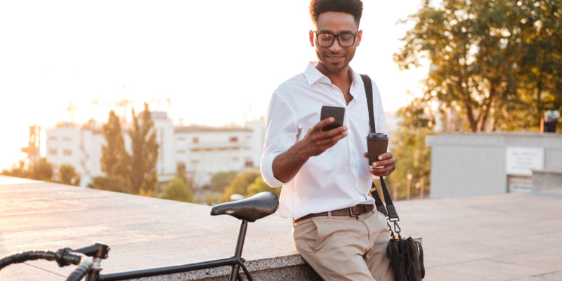 Image of young african man early morning standing near bicycle looking aside chatting by phone outdoors.