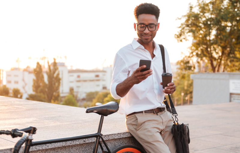 Image of young african man early morning standing near bicycle looking aside chatting by phone outdoors.