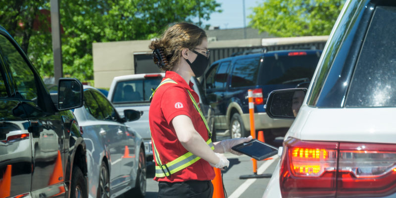 Young youthful fast food worker working at Chick-fil-a drive through amidst cars take orders with tablet.