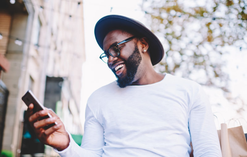 Cheerful African American man in white shirt using mobile phone application for searching cafes near location in downtown, happy dark skinned hipster guy read news from social network walking in city