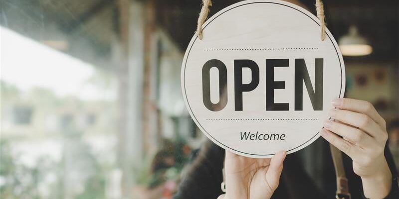 White hand holding an open sign at the front of a business